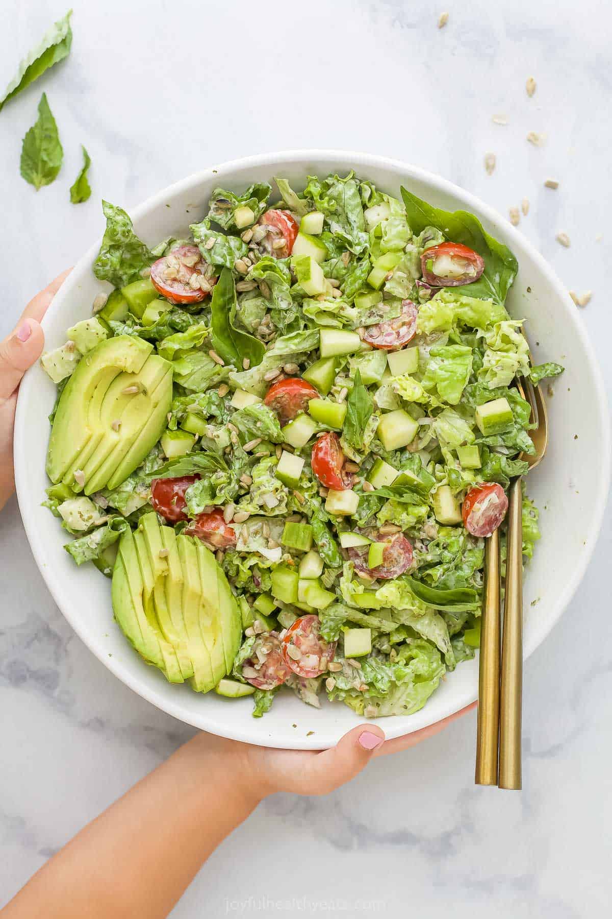 A pair of hands grabbing a big bowl of green goddess salad from a marble countertop