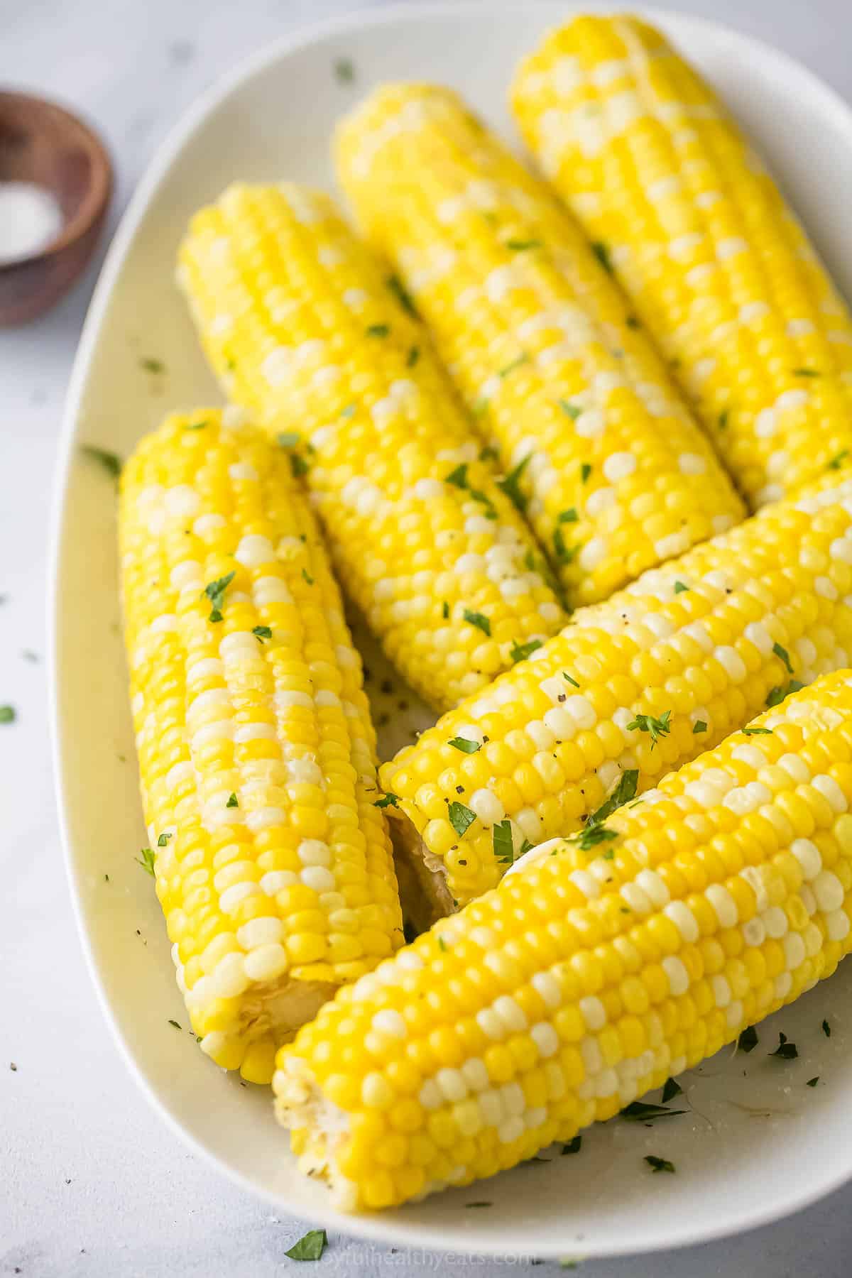 Six cobs of freshly cooked corn on a large plate beside a small dish of salt