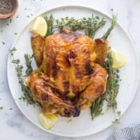 a roasted whole chicken on a platter with fresh herbs and lemon wedges