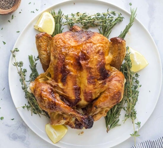 a roasted whole chicken on a platter with fresh herbs and lemon wedges