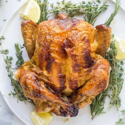 a roasted whole chicken on a platter with fresh herbs and lemon wedges