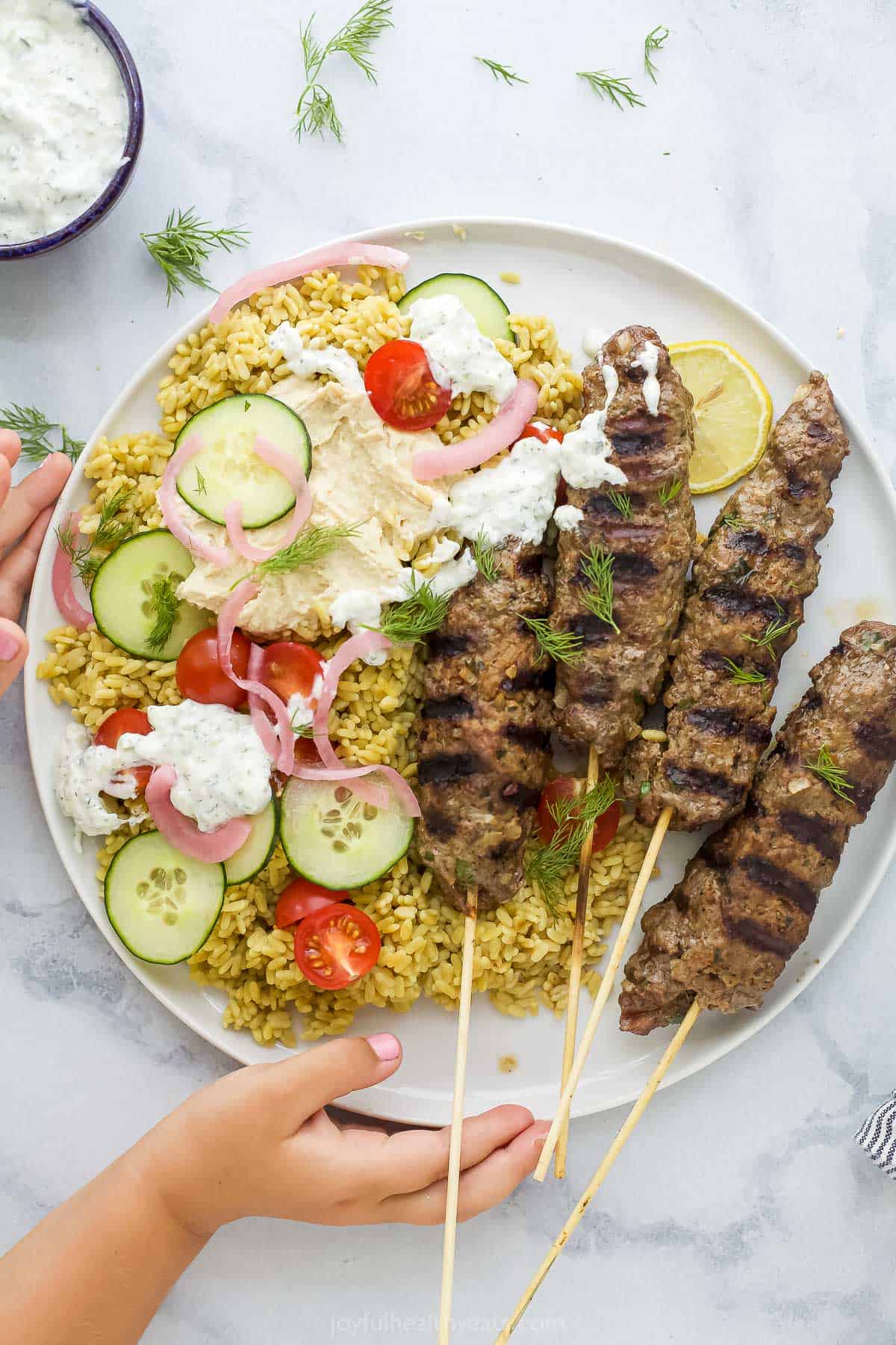 Four seared beef kabobs over rice on a plate with veggies and tzatziki sauce