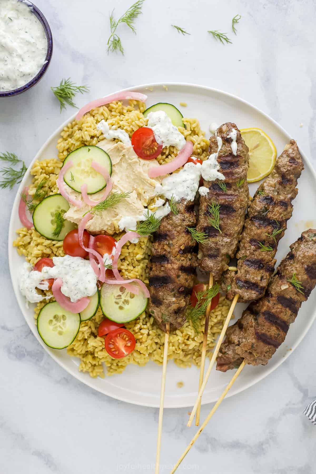 Kefta kabobs on a plate with rice, veggies, tzatziki, pickled onions and fresh chopped herbs