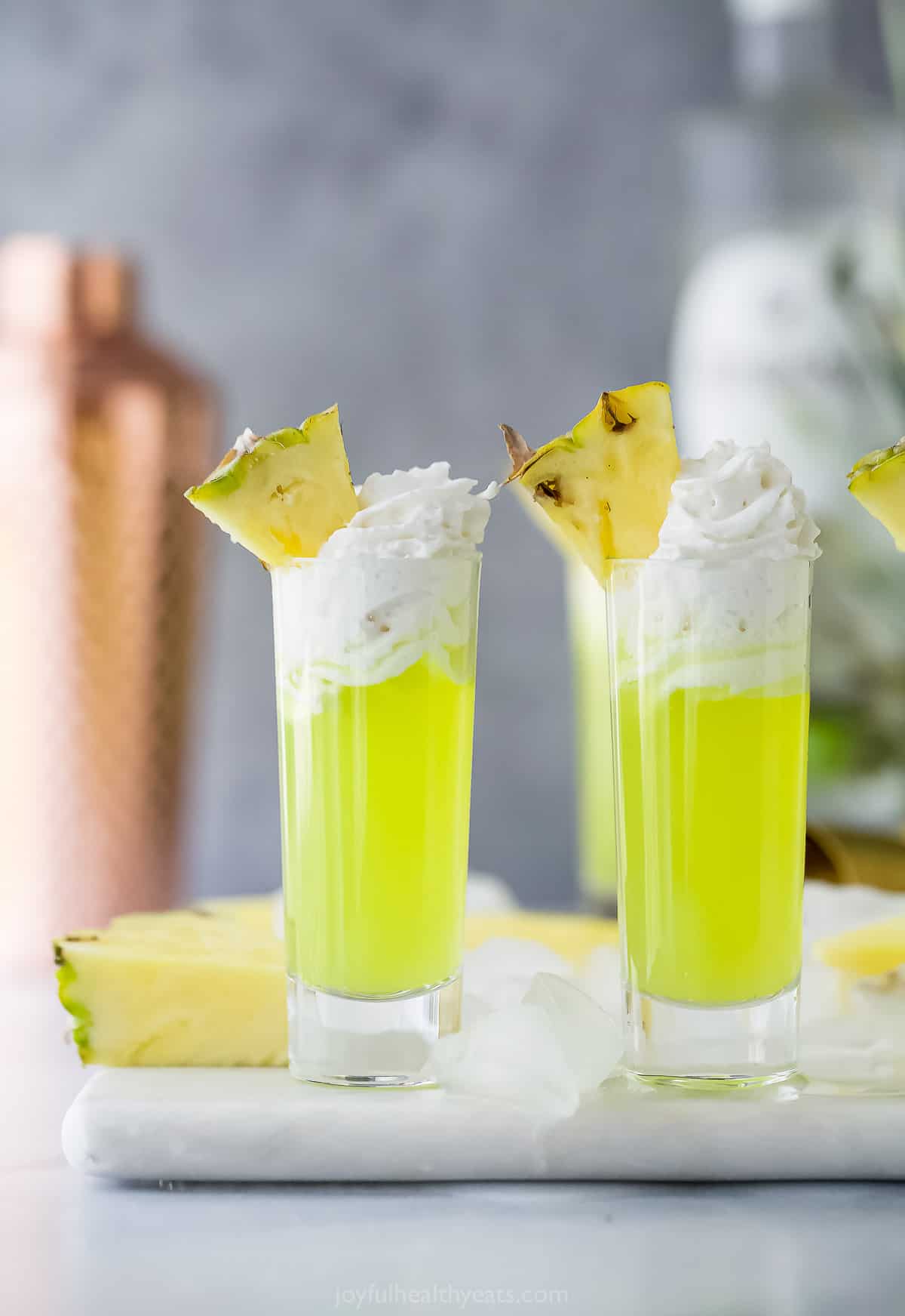 a lime green colored cocktail in a tall shot glsas with whipped cream and pineapple garnish