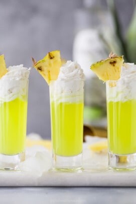 a lime green colored cocktail in a tall shot glsas with whipped cream and pineapple garnish