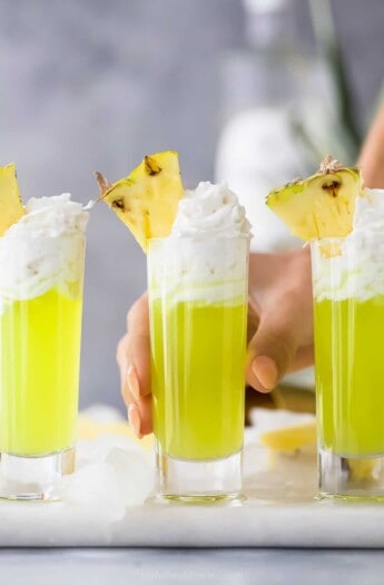 a lime green colored cocktail in a tall shot glsas with whipped cream and pineapple garnish