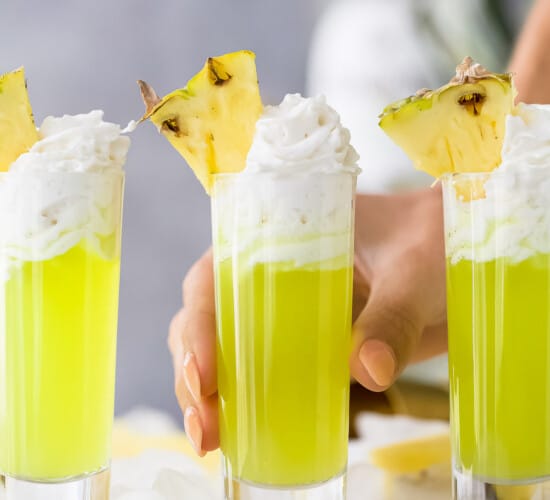 a lime green colored cocktail in a tall shot glsas with whipped cream and pineapple garnish