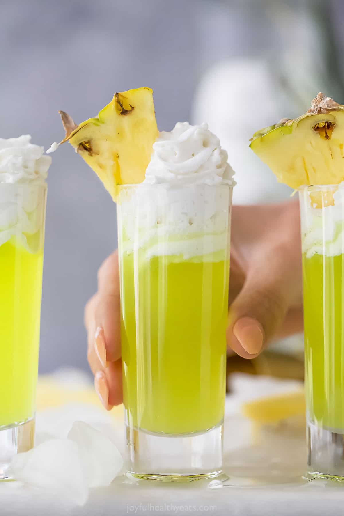 a lime green colored cocktail in a tall shot glsas with whipped cream and pineapple garnish