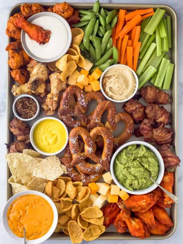 a large sheet tray with an assortment of game day snacks like cheese dip, guacamole, pretzels, chips, cheese cubes, veggies, and chicken wings