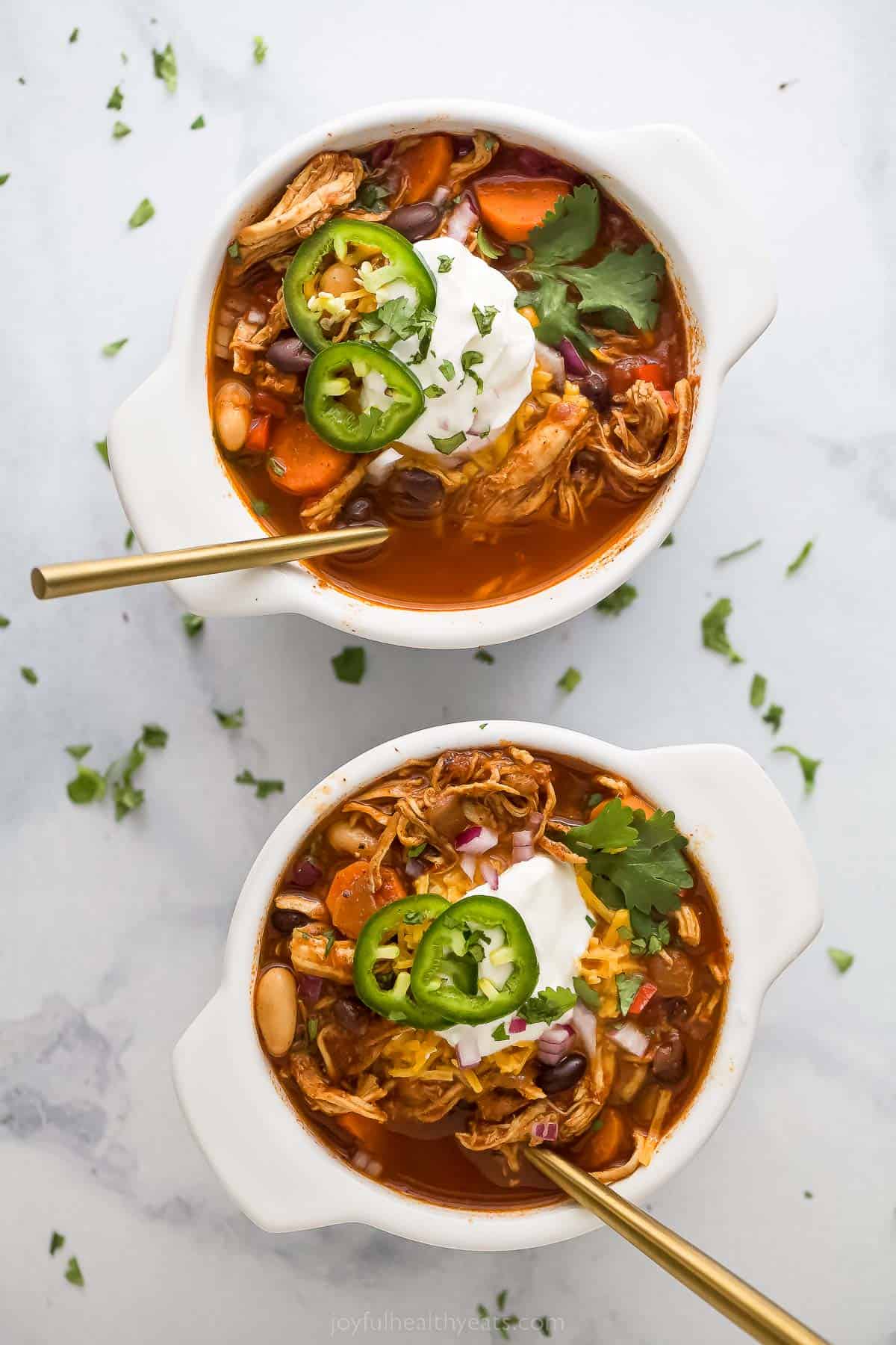 Two bowls of chicken chili with jalapeno, sour cream, and cilantro garnish.