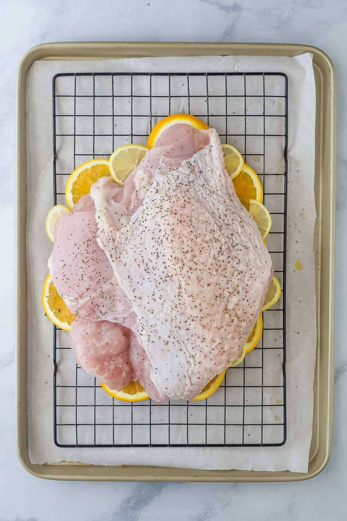 a raw turkey breast on a baking rack seasoned with salt, pepper, and compound butter and laying on a bed of citrus halves
