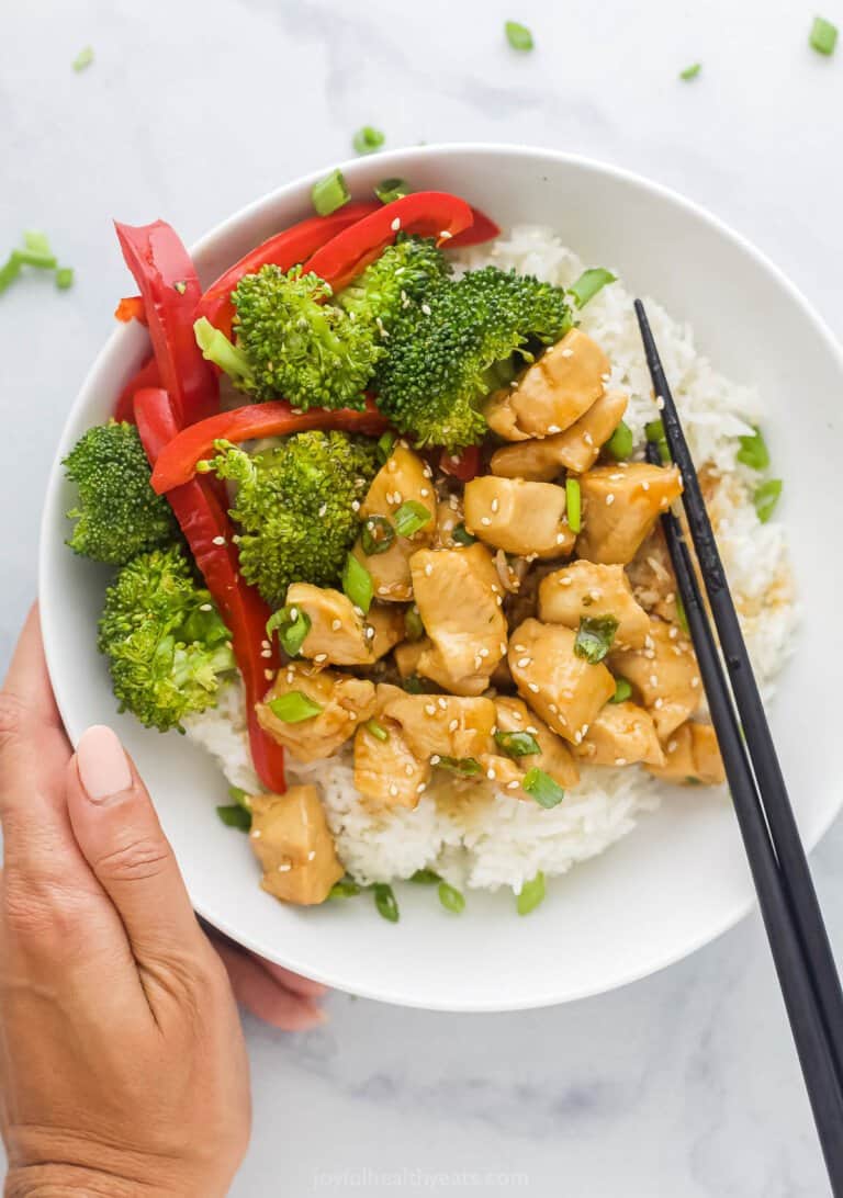 Hand holding a white bowl with teriyaki chicken, veggies and rice.