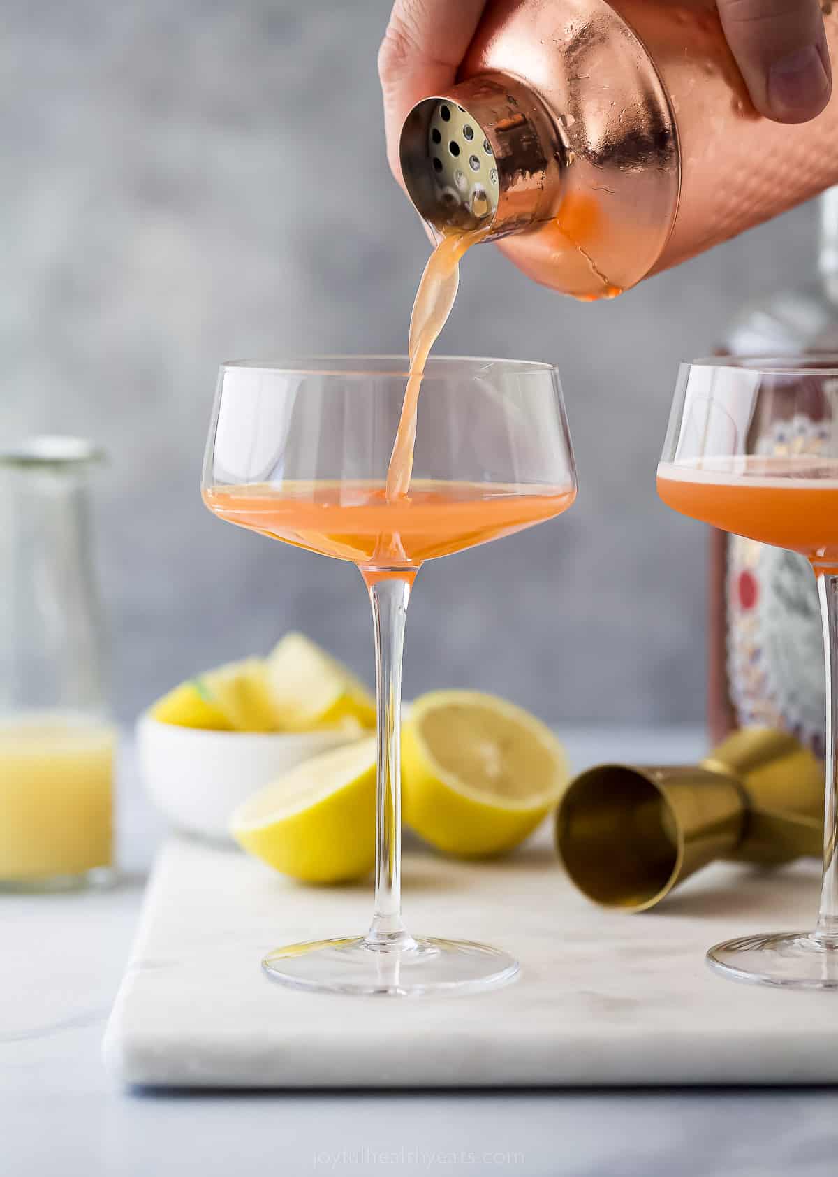 pouring a pale orange cocktail into a tall glass