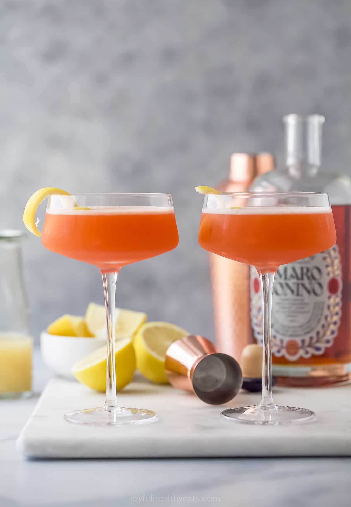 two paper plane cocktails, a simple drink that is pale orange in color, with lemon rind garnish