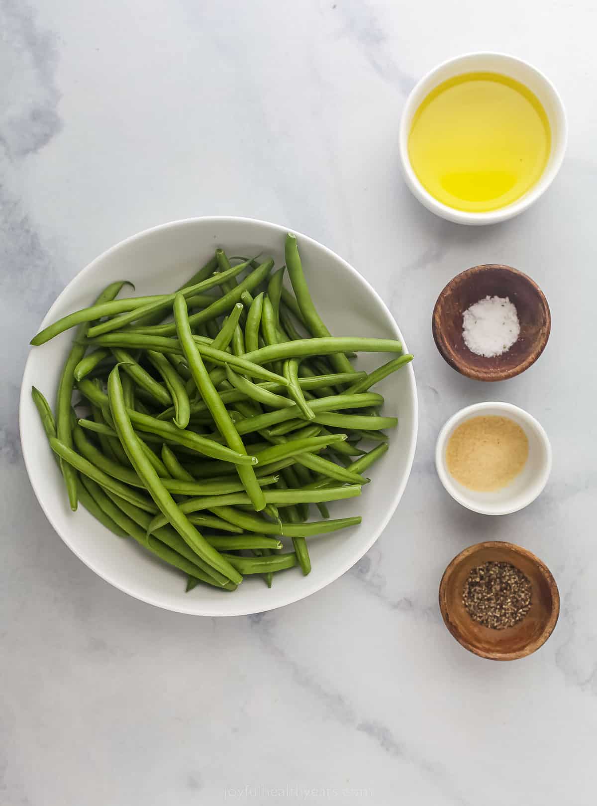 green beans in a bowl alongside small bowls with olive oil, salt, pepper, and garlic powder