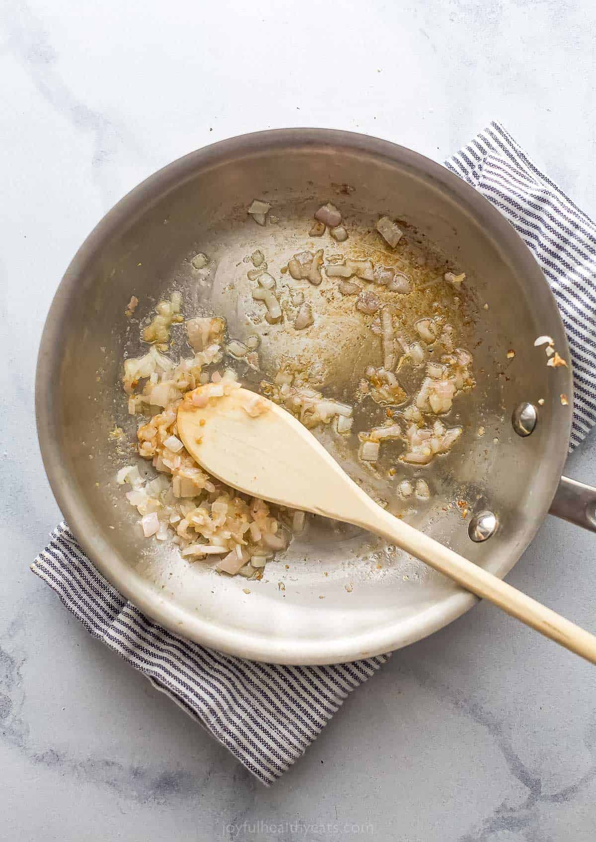 a metal saute pan with cooked shallots and garlic and a wooden spoon