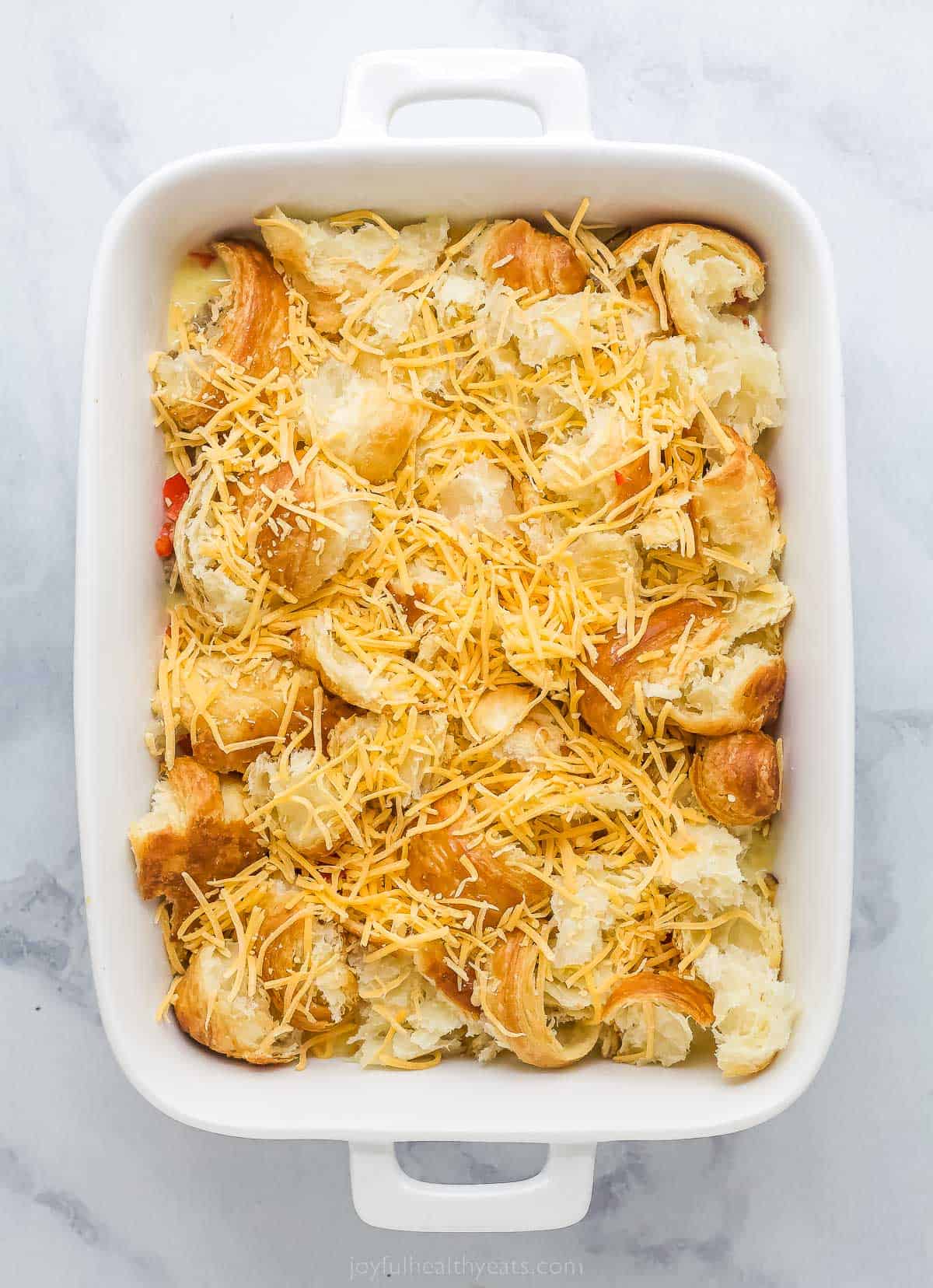 unbaked croissant breakfast casserole with eggs, cheese, peppers, green onions, and cheese in a white ceramic dish