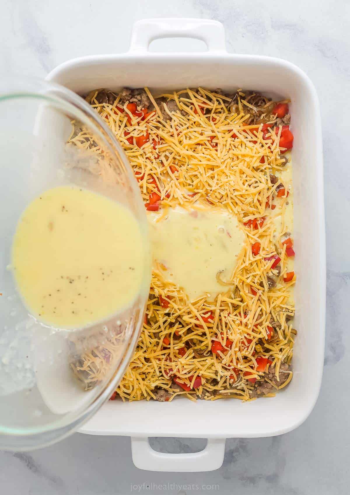 pouring egg into a casserole dish with shredded cheese, sausage, and diced red peppers