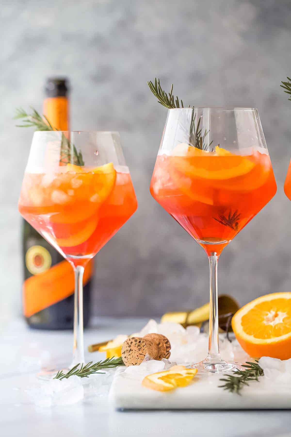 two holiday aperol spritz cocktails - a dark orange drink with orange slices and rosemary garnish