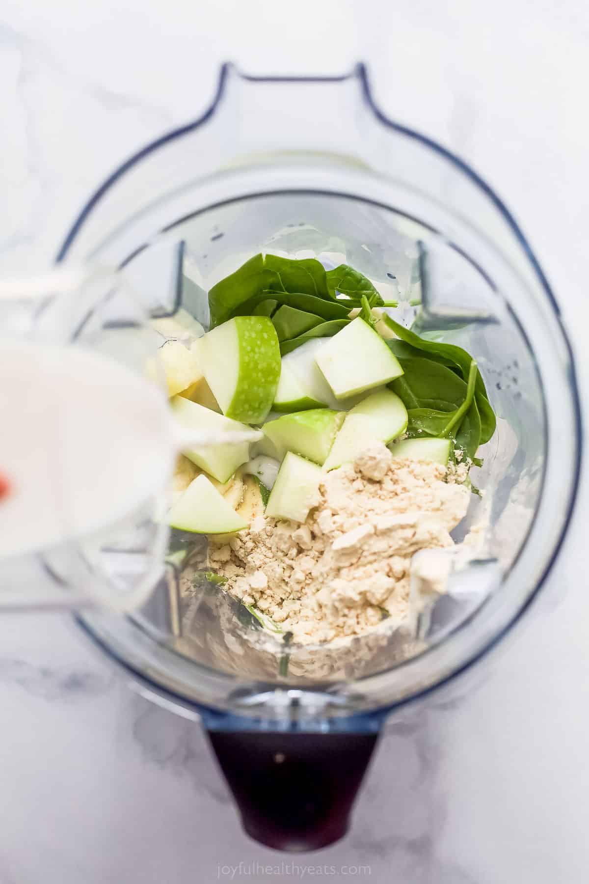 apples, spinach, and protein powder in a blender with milk being poured in