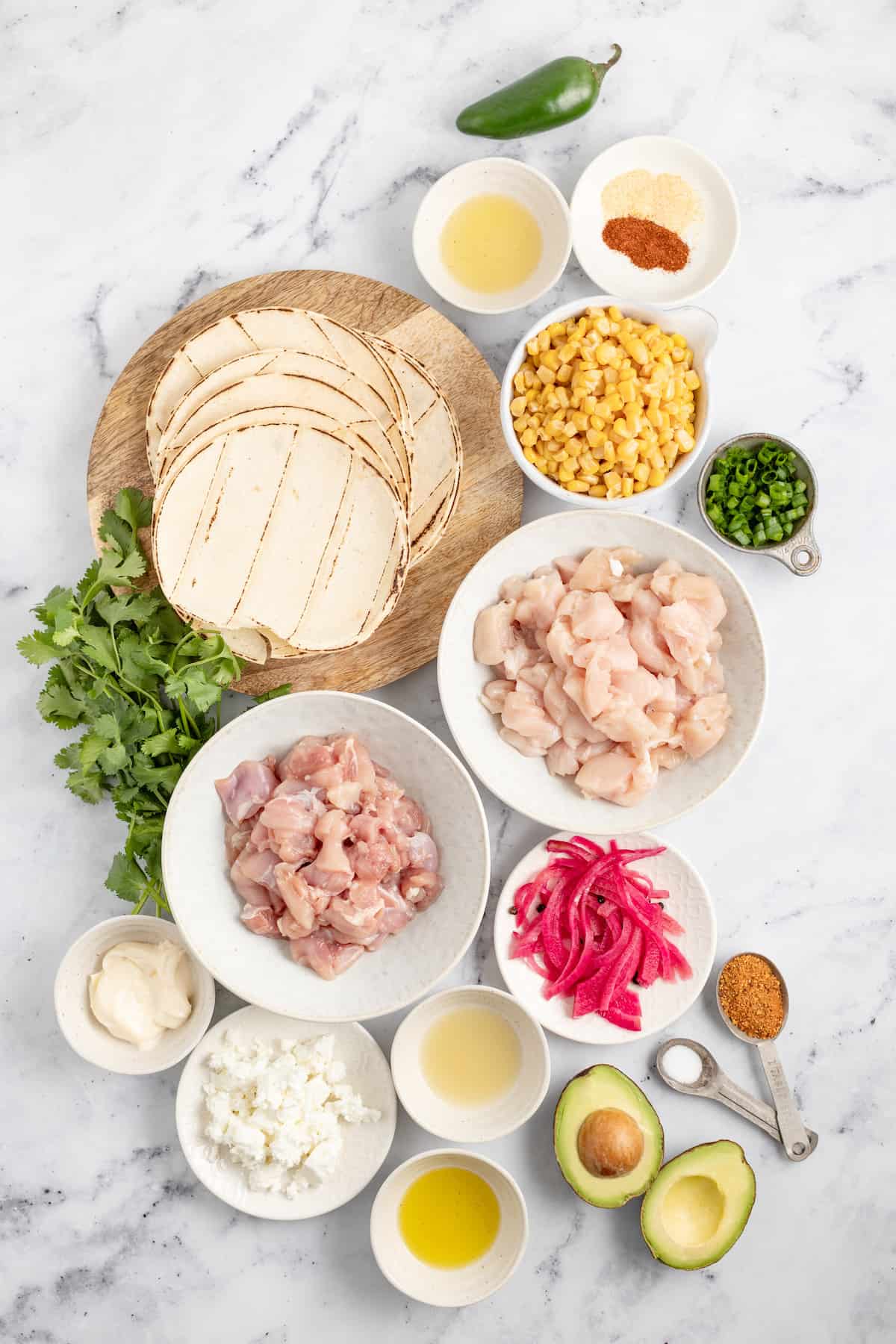 Ingredients to make chicken tacos like tortillas, raw chicken, corn, herbs, pickled onions, and spices