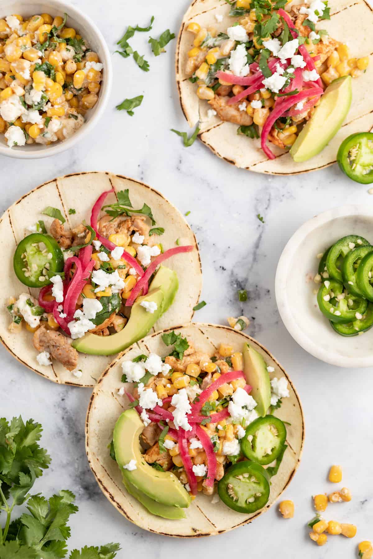 two chicken tacos on a granite counter with pickled onions, chicken, avocado, and corn salad