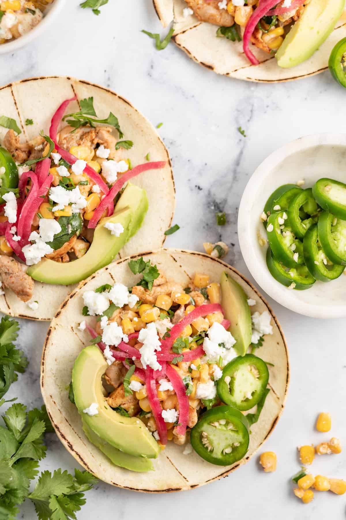 two chicken tacos on a granite counter with pickled onions, chicken, avocado, and corn salad