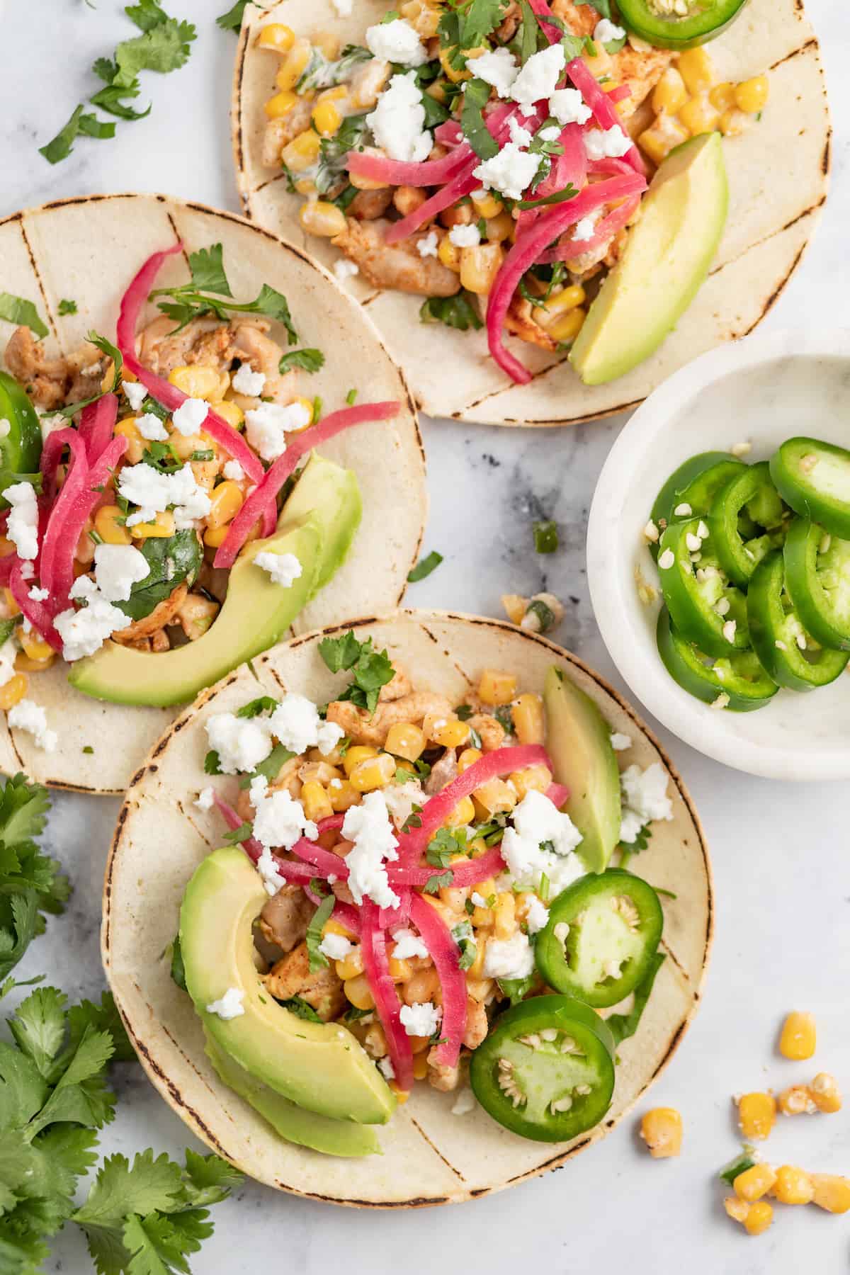 two chicken tacos on a granite counter with pickled onions, chicken, avocado, and corn salad