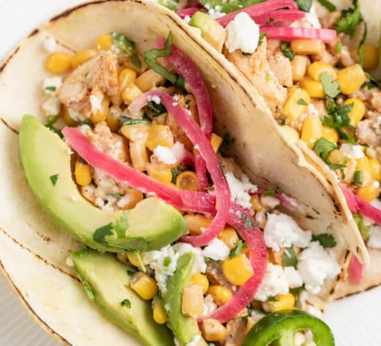 two plated Chicken Street Tacos with avocado, pickled onions, and corn salad