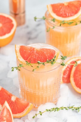 a pale pink beverage known as a greyhiound cocktail in a lowball glass with grapefruit wedge and fresh herb garnish