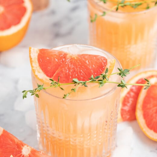 a pale pink beverage known as a greyhiound cocktail in a lowball glass with grapefruit wedge and fresh herb garnish