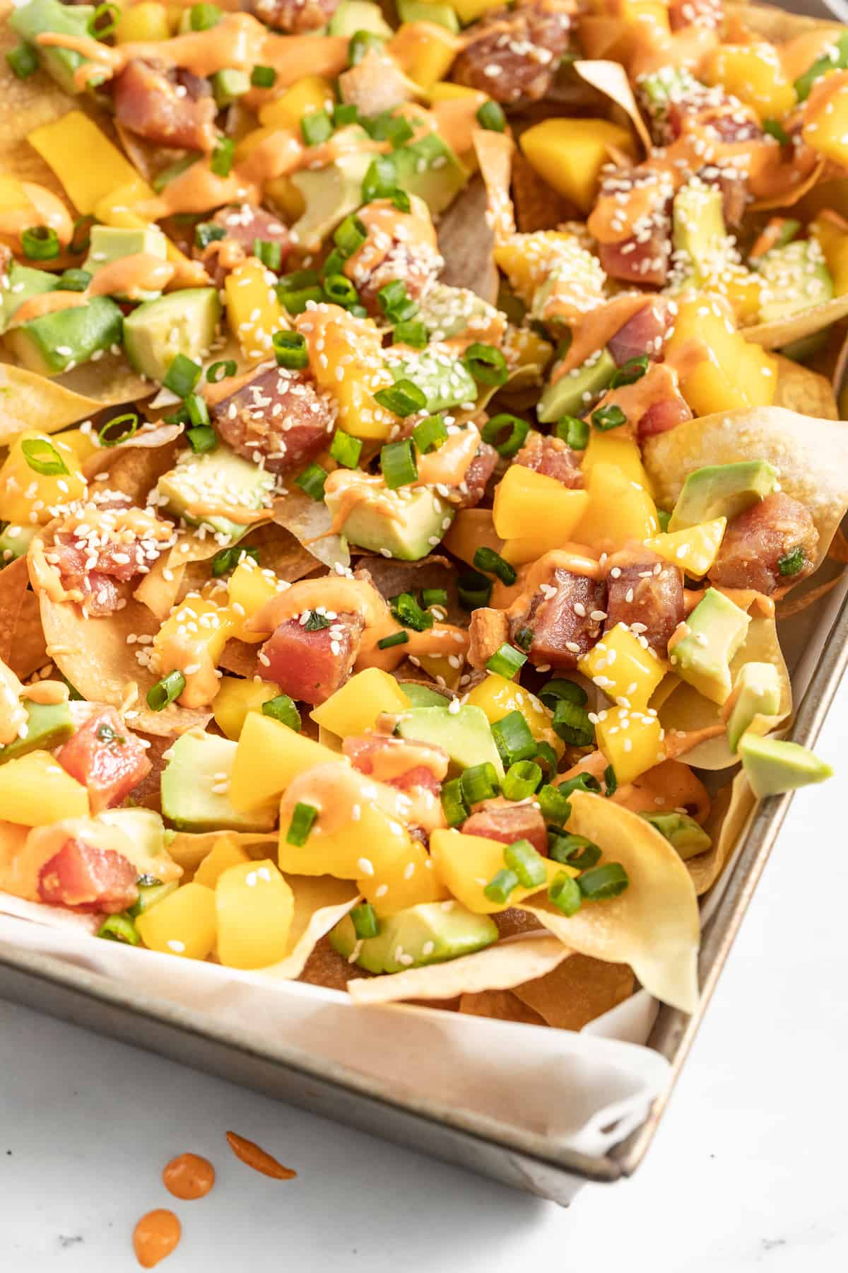 Ahi Tuna Poke Nachos are a unique shareable snack. They are made with homemade wonton chips, seasoned ahi tuna, mango, and avocado. a platter of wonton chips topped with diced ahi tuna, mango, scallions, and sriracha mayo