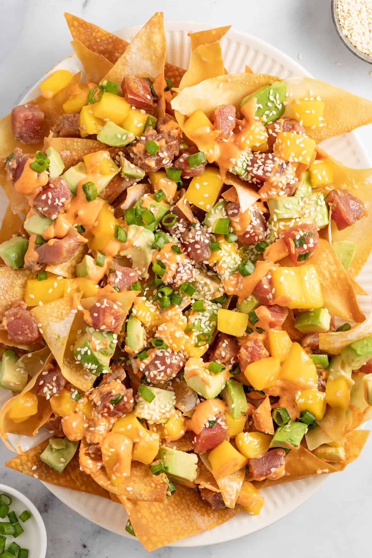 Ahi Tuna Poke Nachos are a unique shareable snack. They are made with homemade wonton chips, seasoned ahi tuna, mango, and avocado. a plate of ahi tuna poke nachos with diced mango, green onions, and sriracha mayo