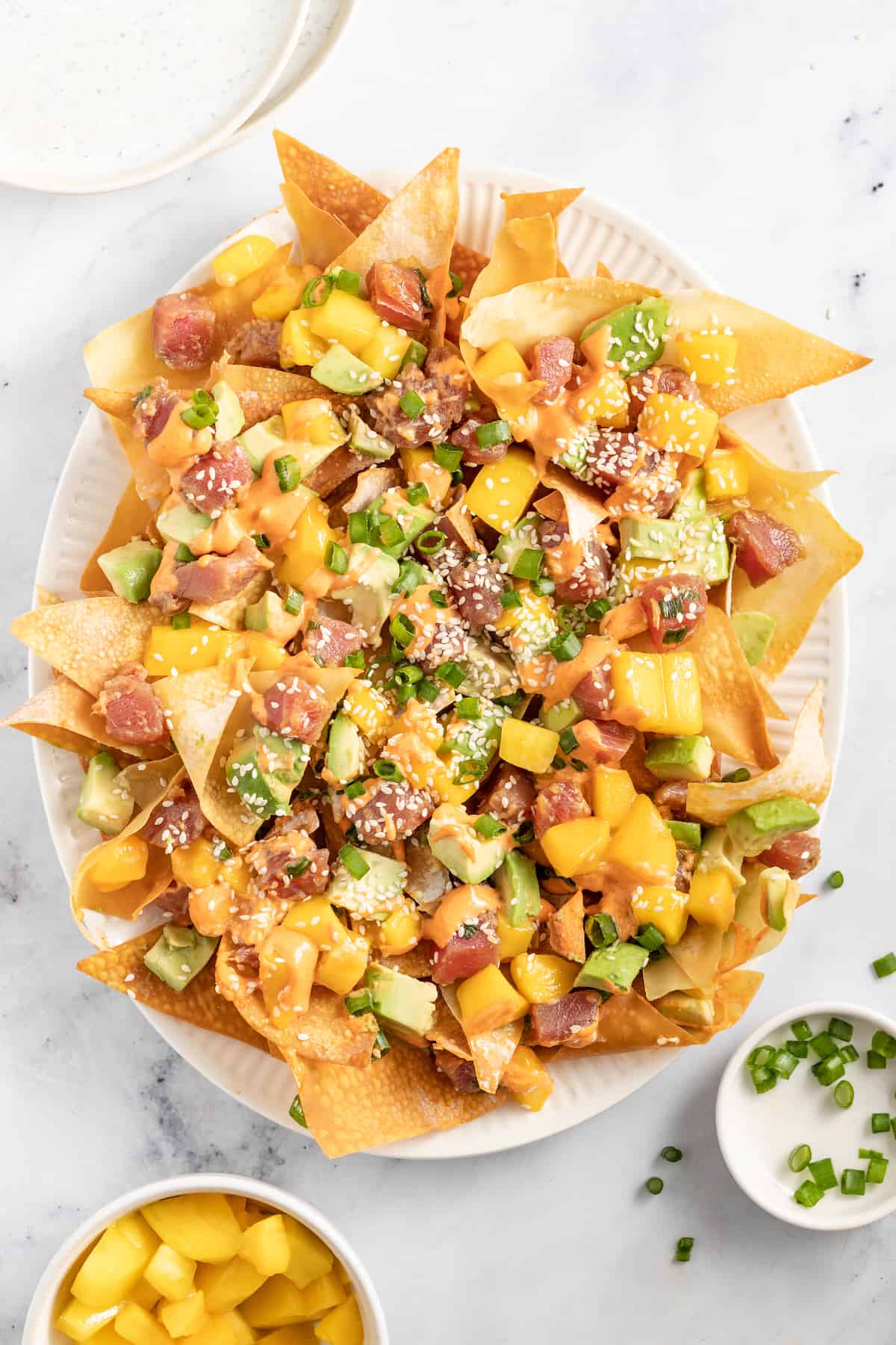 Ahi Tuna Poke Nachos are a unique shareable snack. They are made with homemade wonton chips, seasoned ahi tuna, mango, and avocado. a plate of ahi tuna poke nachos with diced mango, green onions, and sriracha mayo