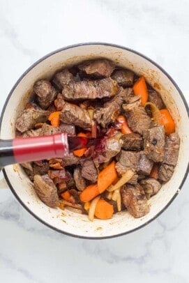 pouring wine over chunks of beef with carrots and onions in a dutch oven