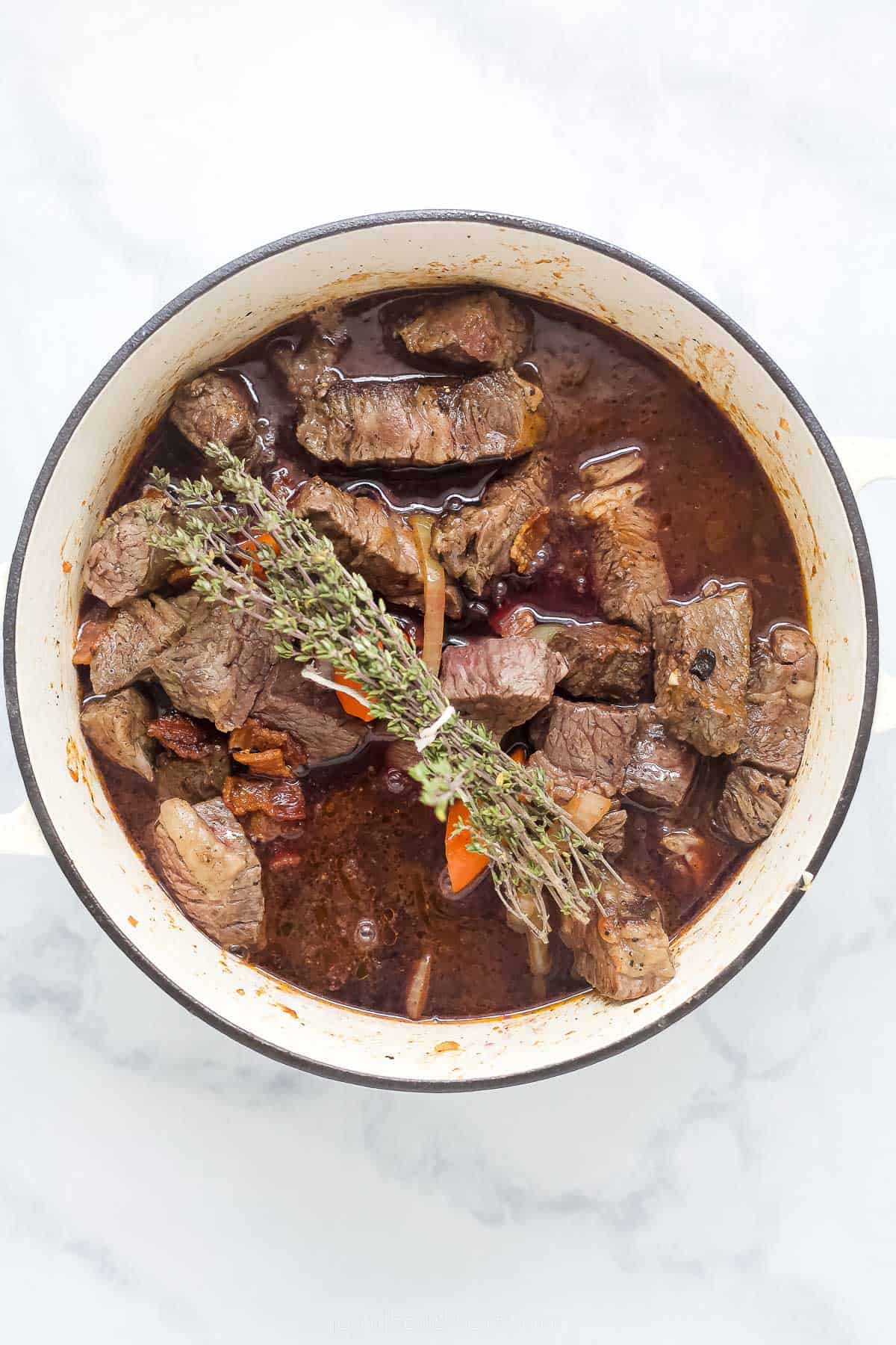 chunks of beef in broth with a herb bundle in a dutch oven