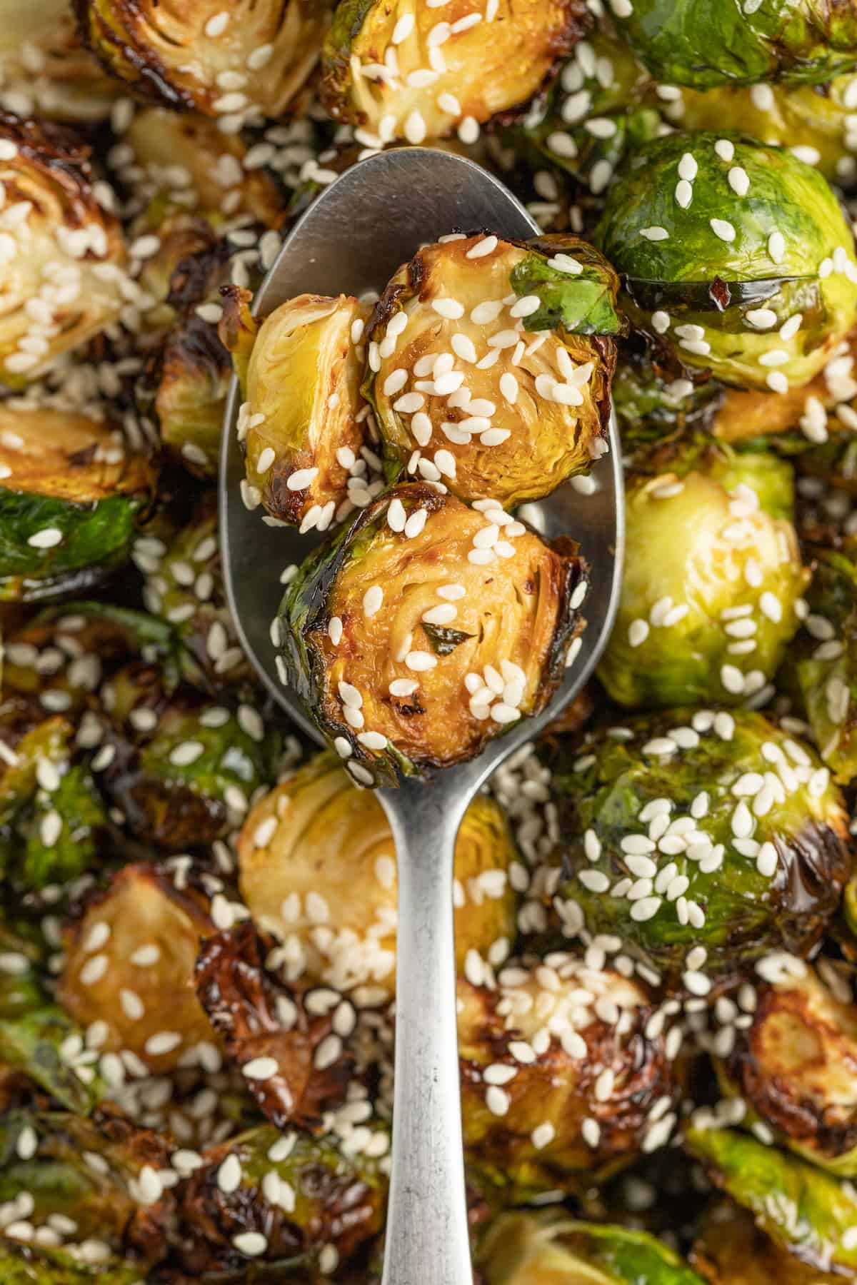 These Crispy Asian Air Fryer Brussels Sprouts are crunchy little bites filled with lots of soy, ginger, garlic, and spicy flavor. a spoon holding brussels sprouts that have been fried over a bowl of more brussels sprouts