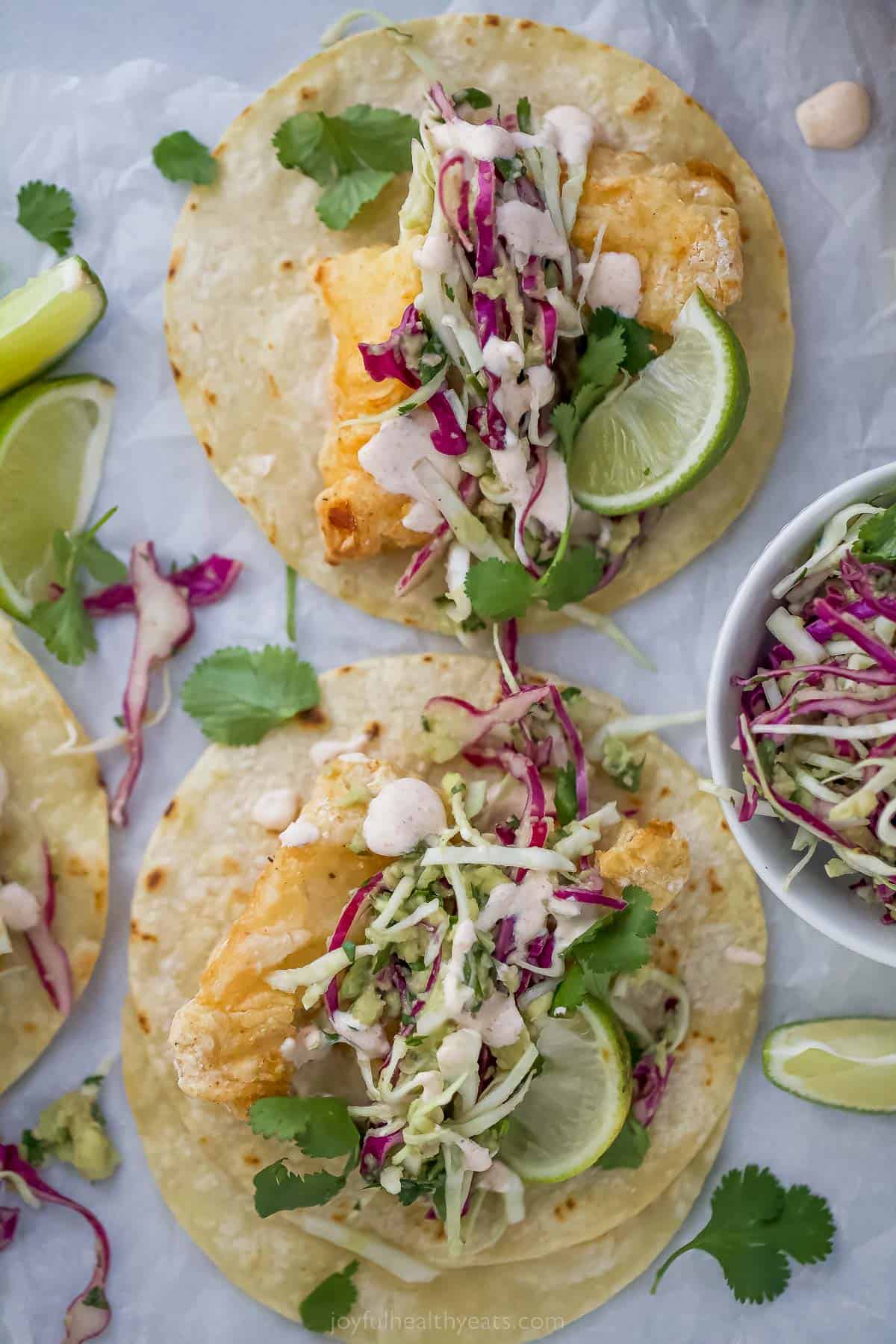 Beer Battered Air Fryer Fish Tacos are dipped in a tasty batter and fried the healthy way! Add slaw and Chipotle Crema for the perfect taco! top view of air fryer beer battered fish tacos with slaw and lime wedge as a garnish