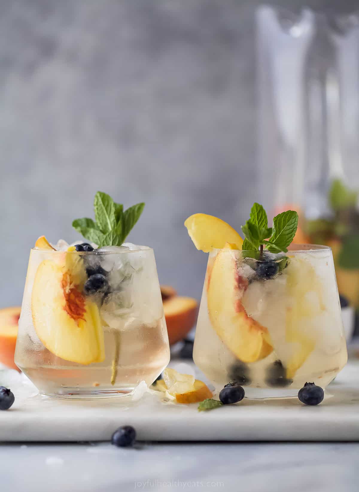 This icy peach sangria with blueberries, peach Schnapps, and mint is the fruity cocktail you'll be sipping on all summer. Two peach sangria glasses side by side.