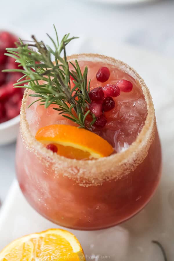 Close-up of the pomegranate cocktail with an orange slice, pomegranate arils, and a rosemary sprig as garnishes.