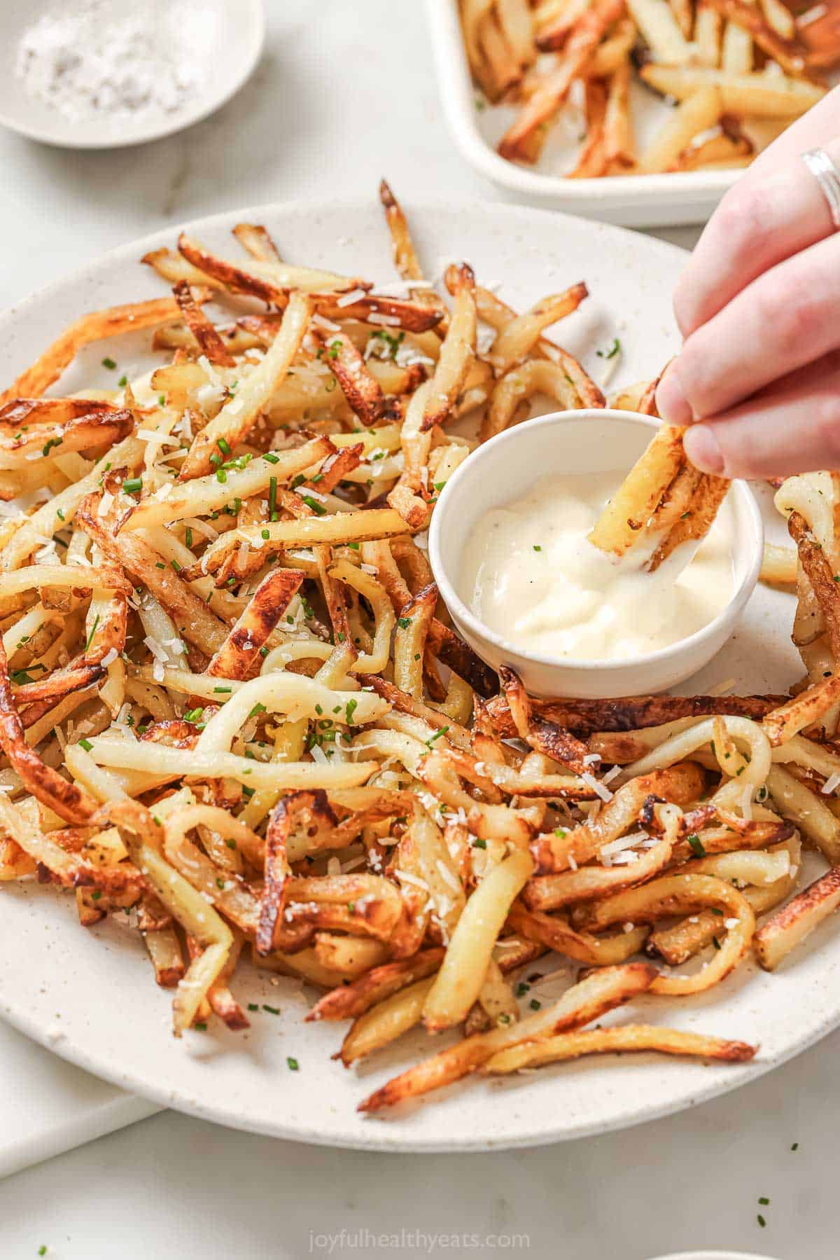 Cheesy Parmesan Truffle Fries are the perfect snack or party app when you’re feeling a little fancy. Baked in the oven and super crispy! Overhead view of baked Parmesan truffle fries on platter, with hand dipping fries into aioli