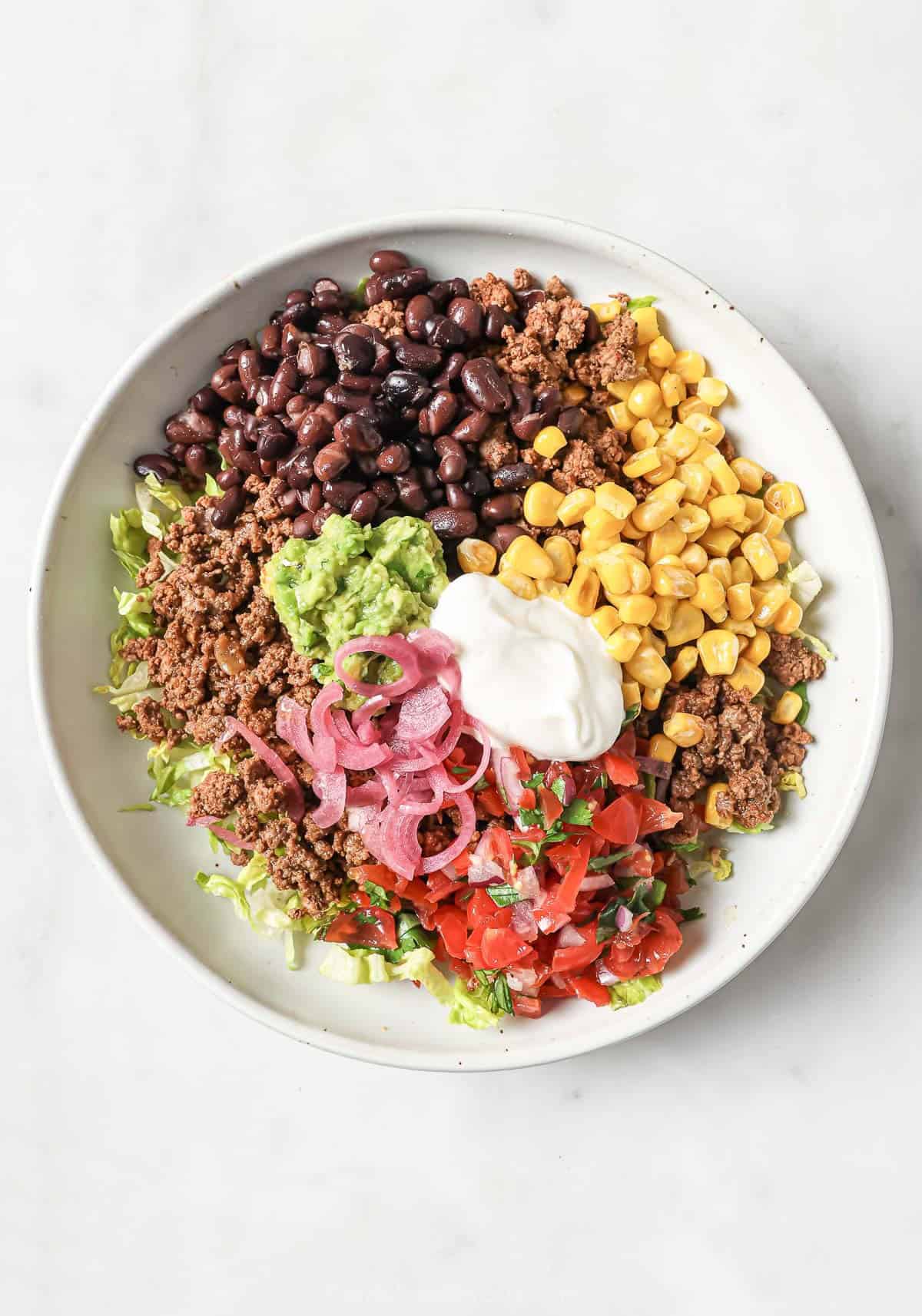 Overhead view of taco salad bowl with corn, beans, ground beef, guacamole, and sour cream over lettuce