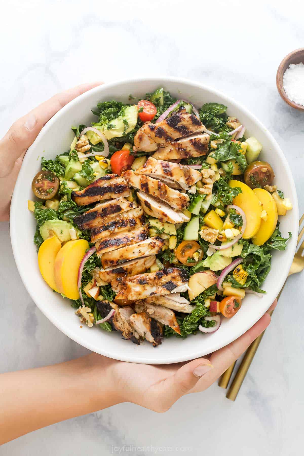 With sweet corn, peaches, tomatoes, grilled chicken and honey mustard dressing, this summer salad recipe is the perfect patio dinner! Hands holding summer salad with grilled chicken