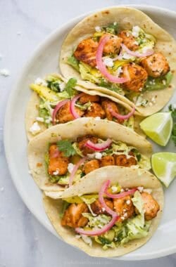 Four blackened salmon tacos on plate with lime wedges.
