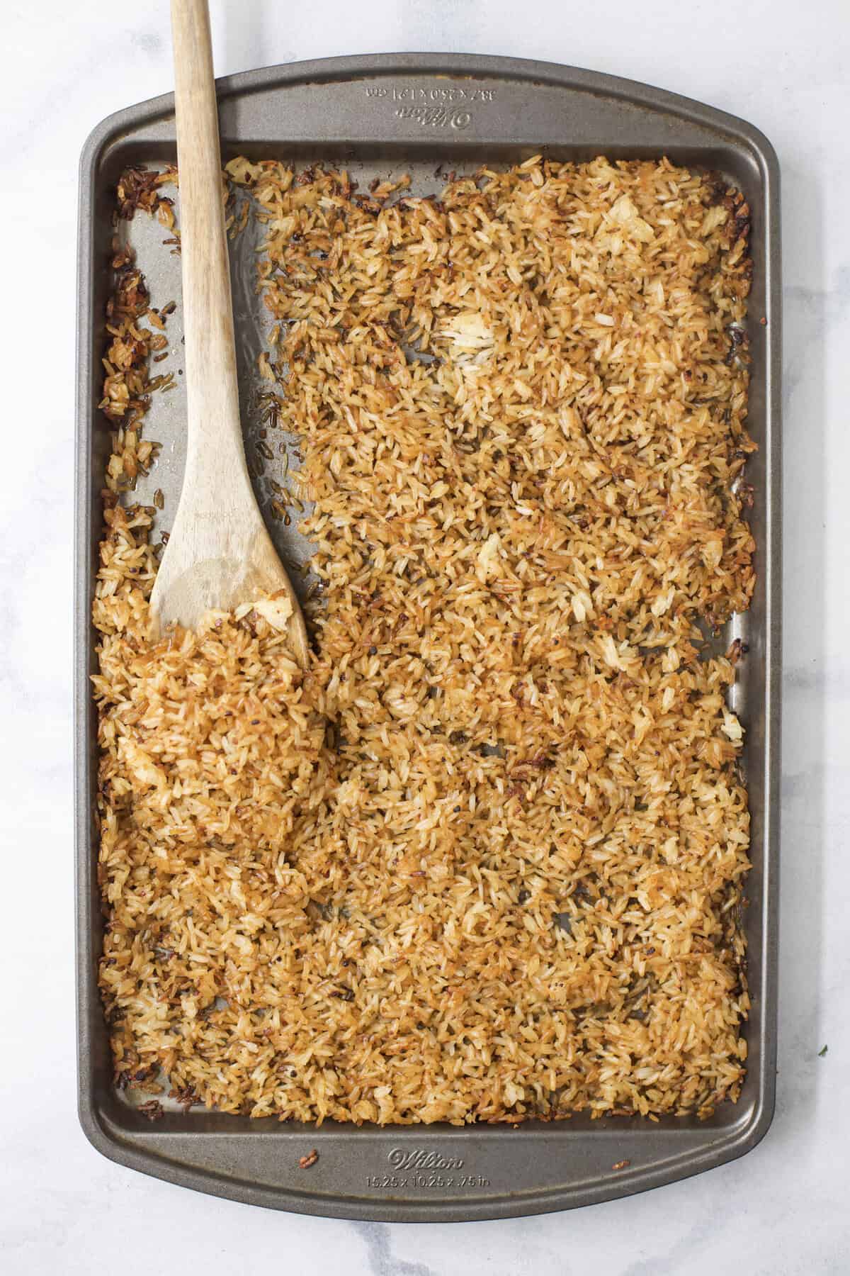 This viral TikTok sheet pan crispy rice is tossed in savory sauce and baked until golden for the perfect addition to salads, bowls and more! Wooden spoon on pan of crispy rice.