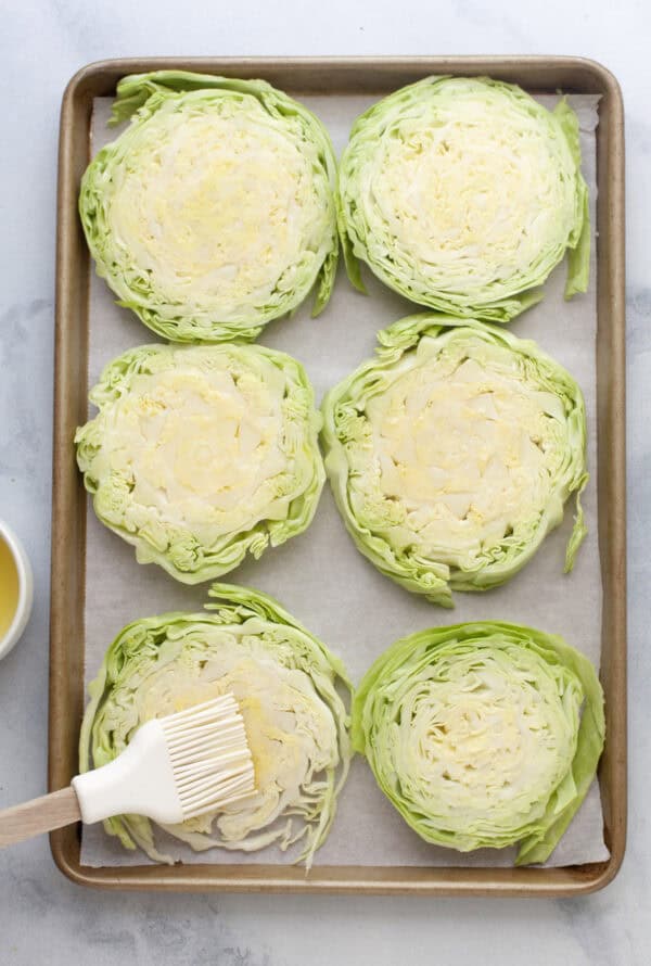 Six cabbage steaks on sheet pan with one steak being brushed with oil.