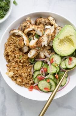 Spoon in bowl with sliced chicken, crispy rice, cucumber salad, and avocado.