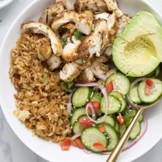 Spoon in bowl with sliced chicken, crispy rice, cucumber salad, and avocado.