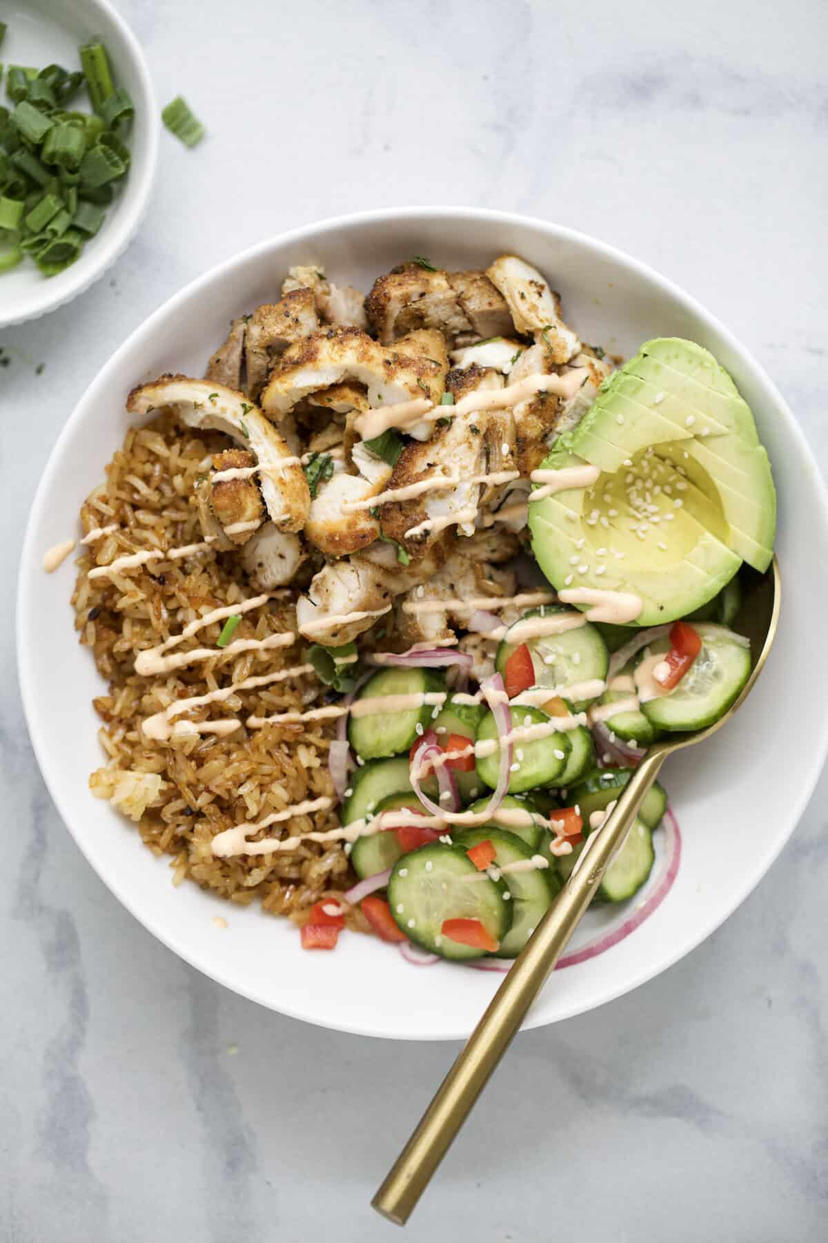 Spoon in bowl with crispy rice, air fried chicken thighs, avocado, and cucumber salad.