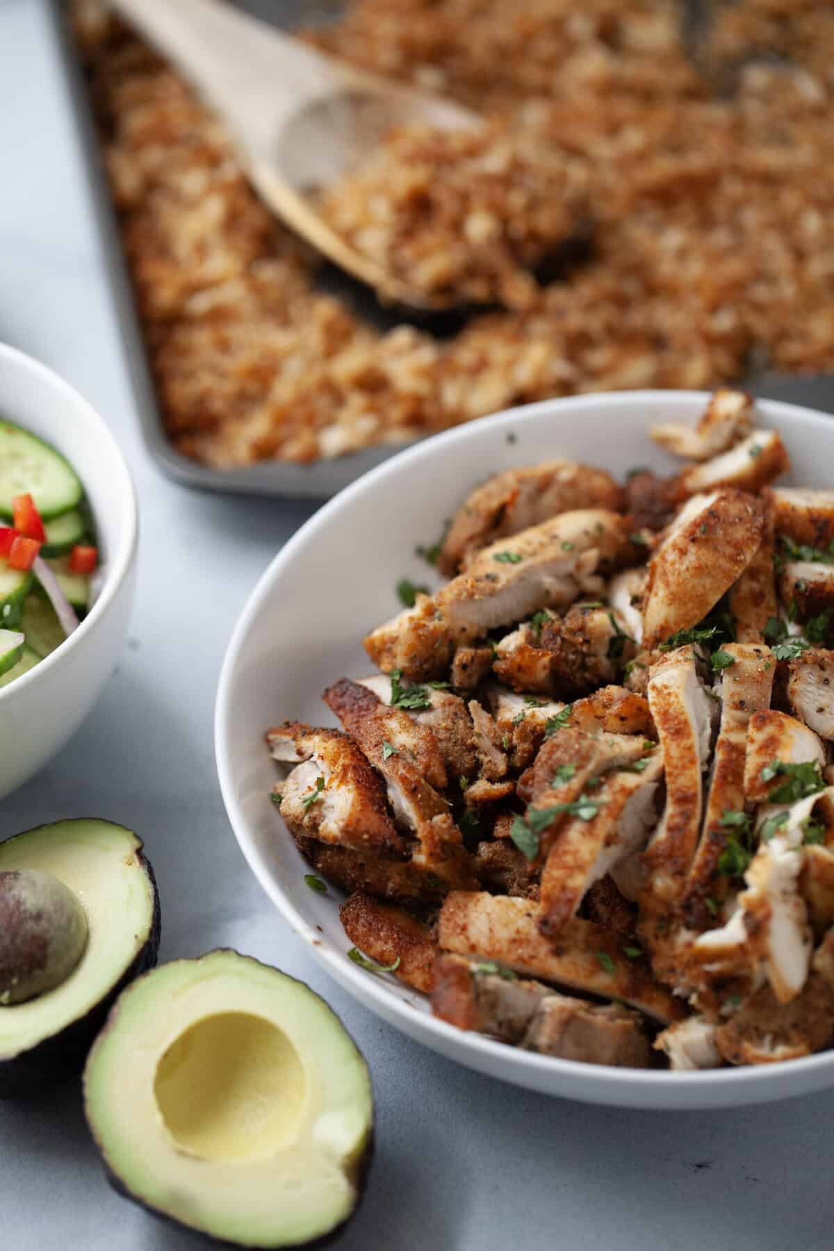 Bowl of sliced crispy chicken thighs set next to halved avocado.
